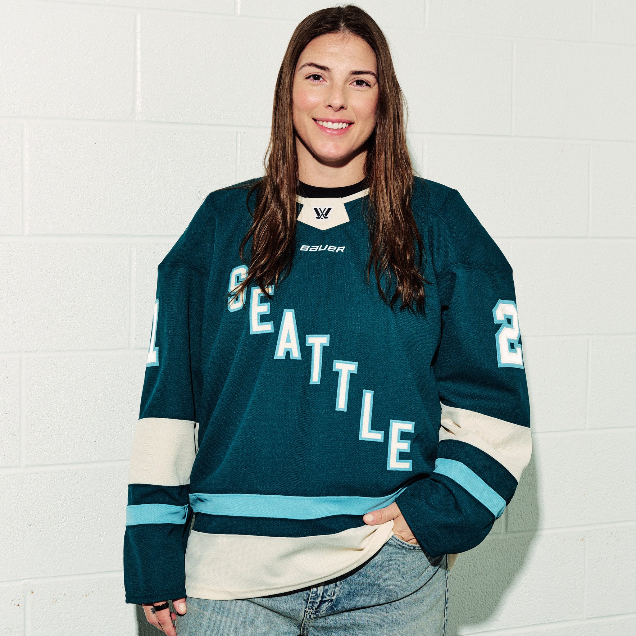 Hilary Knight wearing her green PWHL Seattle jersey with, standing against a white tiled wall with her hand in her pocket.