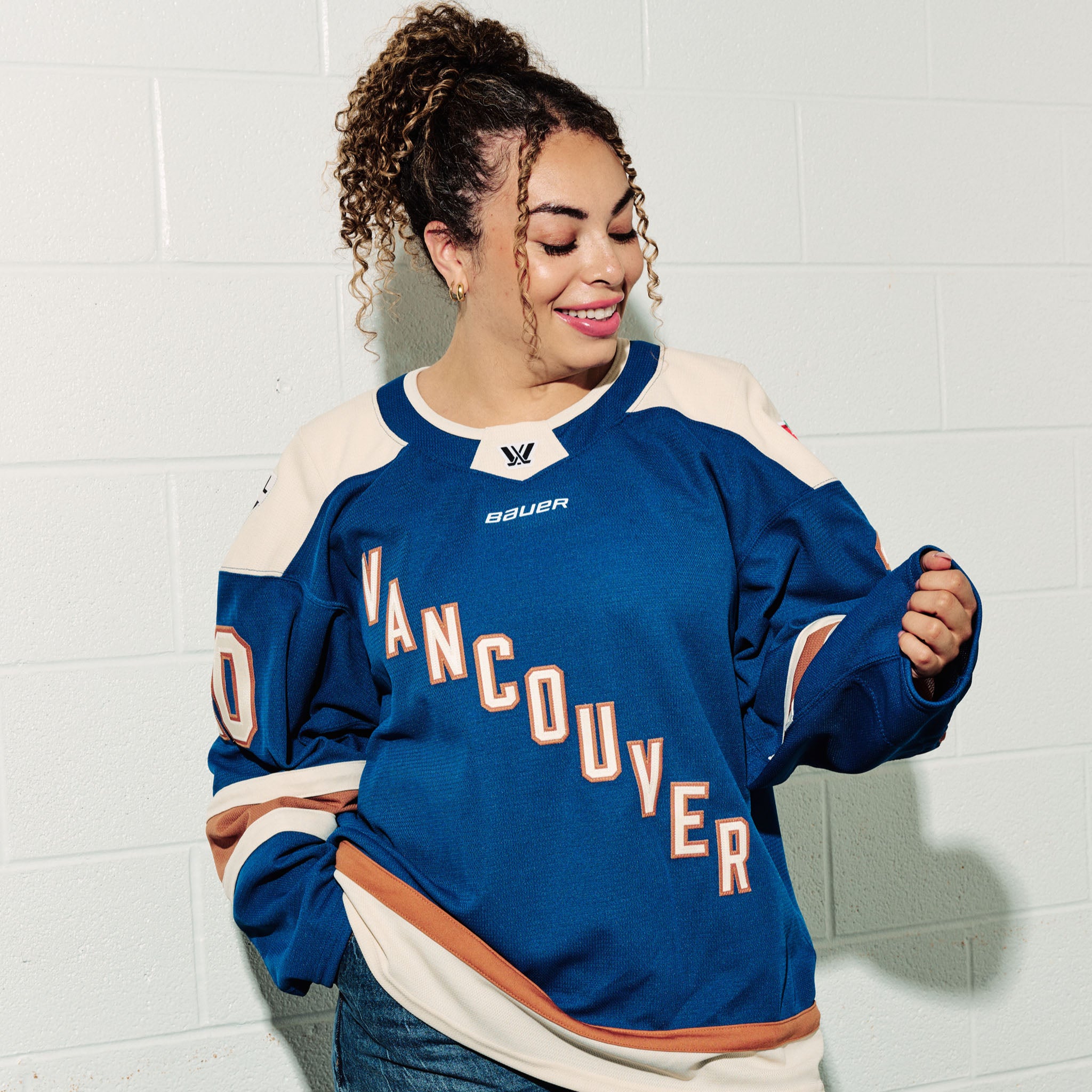 Sarah Nurse wearing her blue PWHL Vancouver jersey with, standing against a white tiled wall looking at her hand.