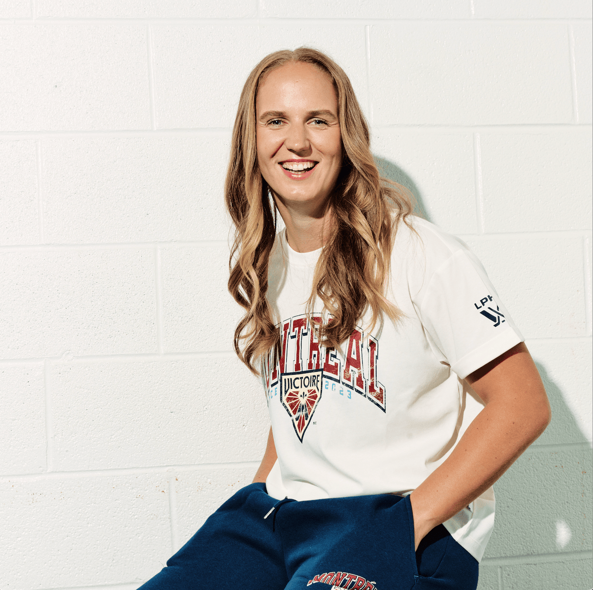 Person wearing a white t-shirt with a logo and text, sitting against a light-colored wall.