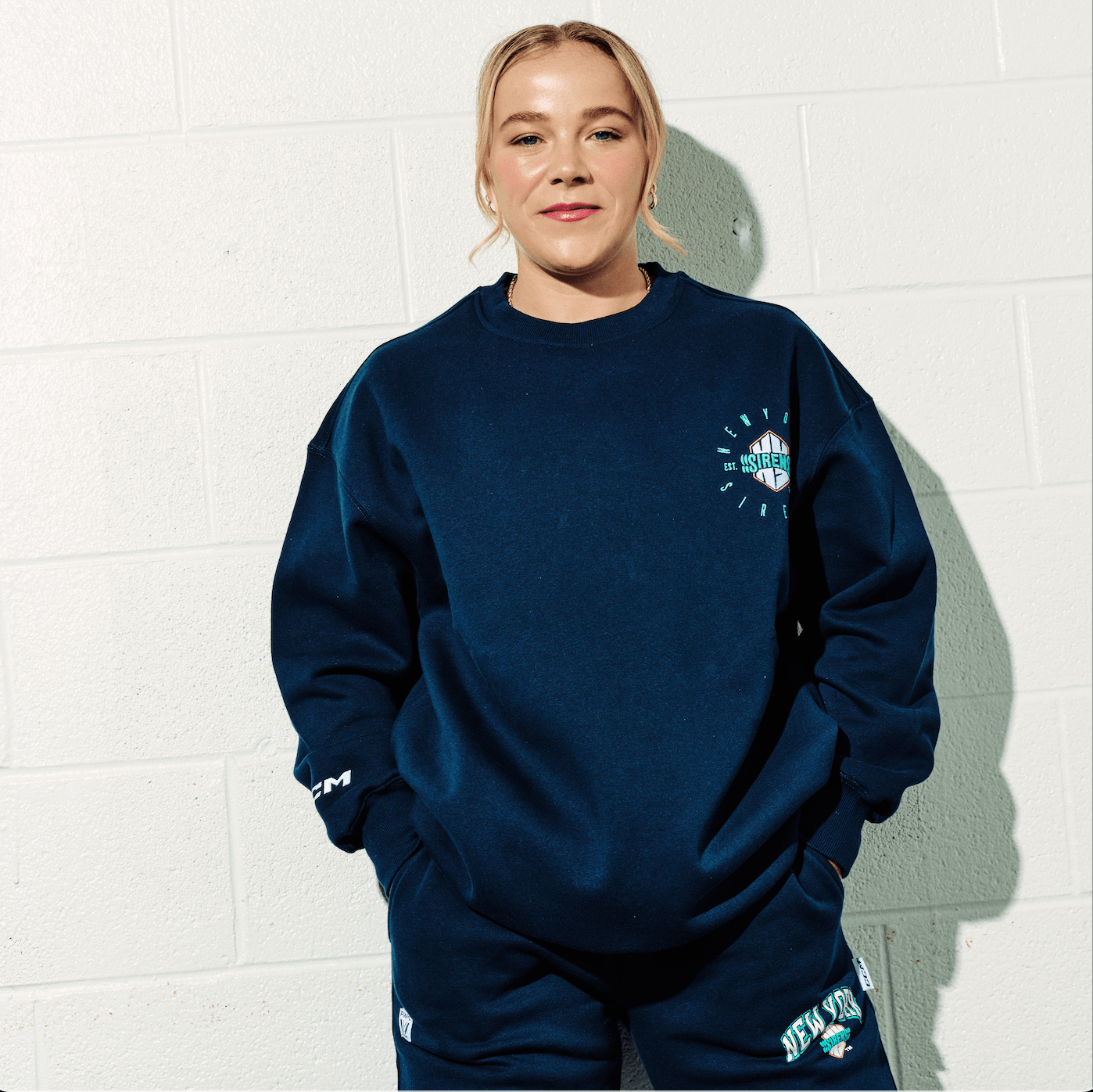 Person wearing a navy blue sweatshirt with a logo against a white brick wall