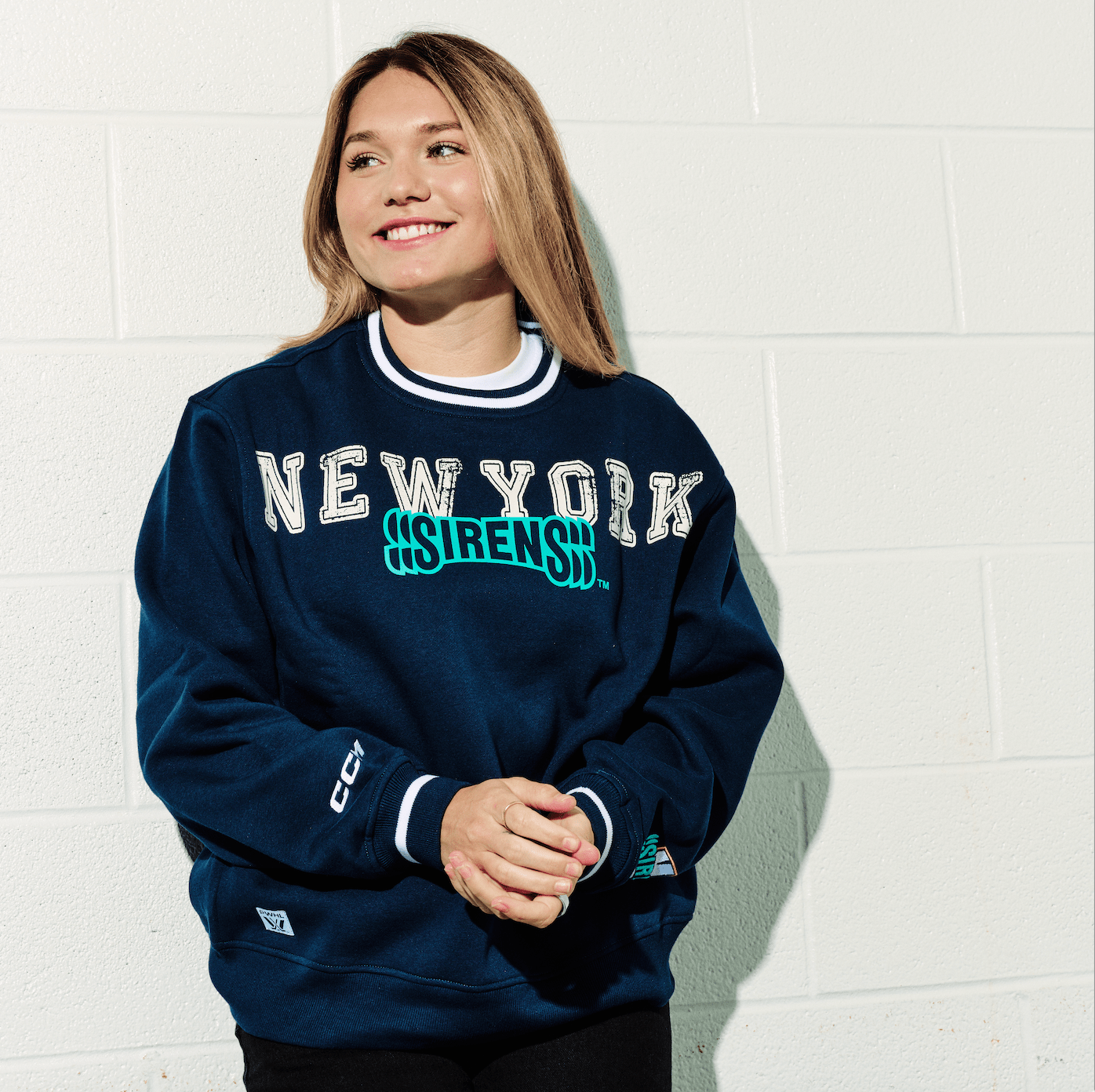 Person wearing a navy blue sweatshirt with 'New York Sirens' text against a white brick wall.