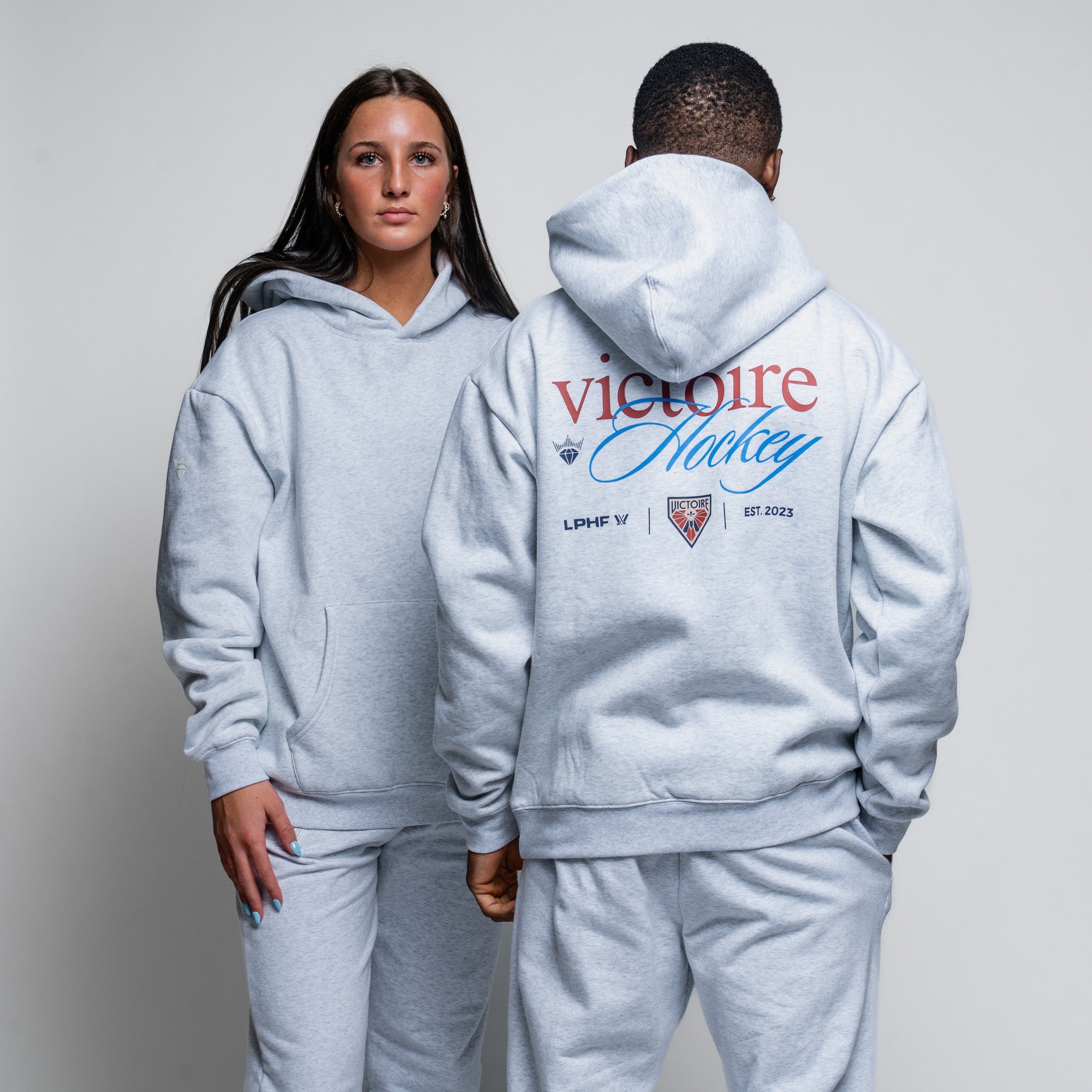 Two people wearing matching gray hoodies with visible branding on a plain background