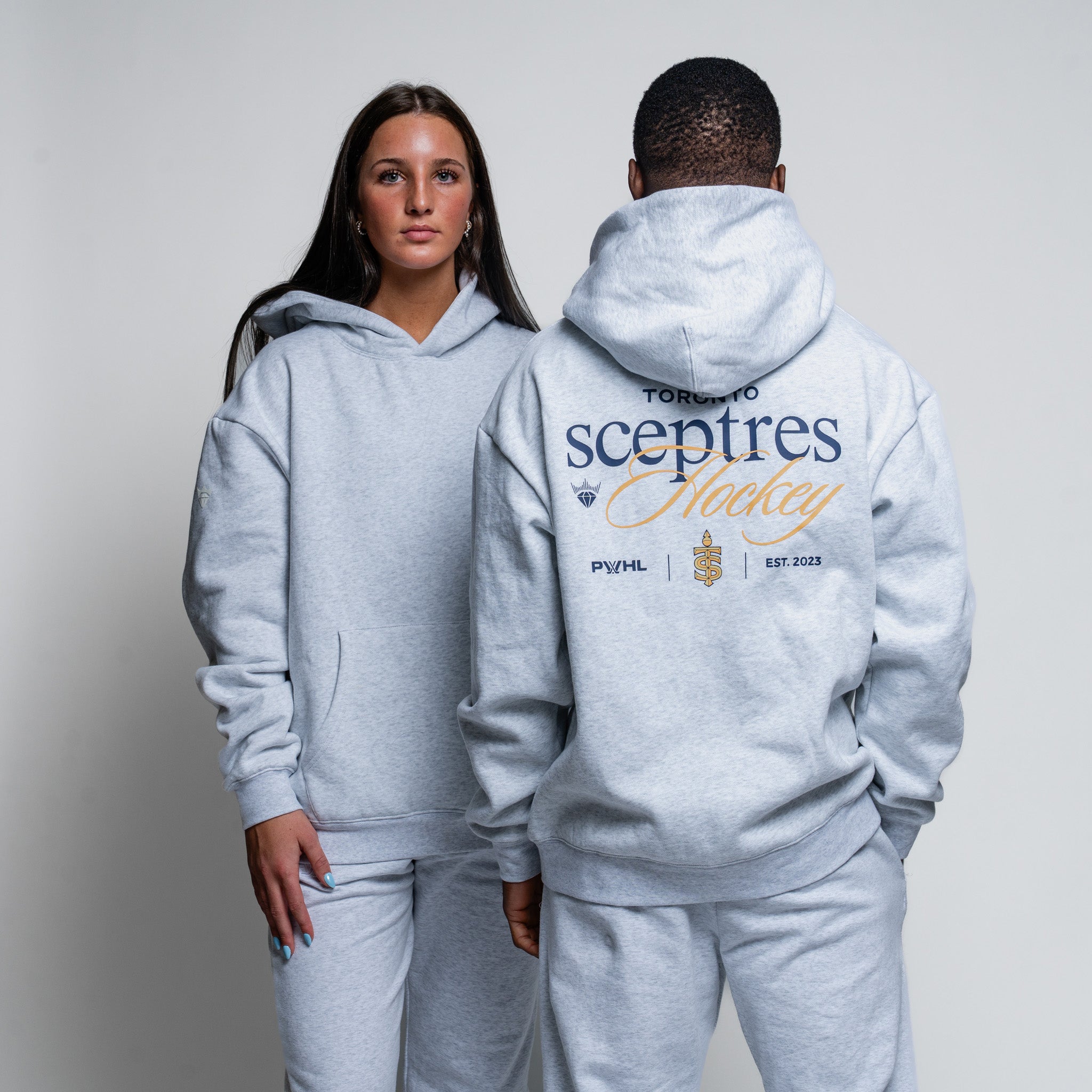 Two people wearing matching gray hoodies with branding on a plain background