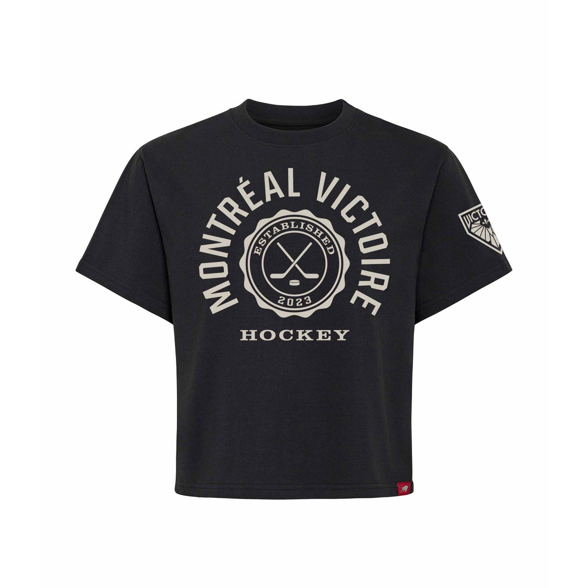 Front of a black short sleeve shirt. 'MONTREAL VICTOIRE' is written in a white arch of text around a circular emblem with crossed hockey sticks and 'ESTABLISHED 2023' inside with 'HOCKEY' below it. A white mono-color Montreal Victoire logo is on the left sleeve.