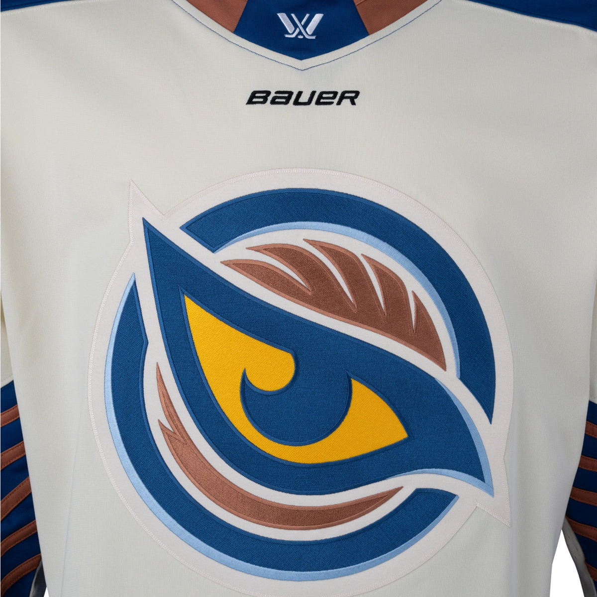 Front of a cream-colored jersey with the Vancouver Goldeneyes logo in the center. The tan collar has a blue color block with the PWHL logo and the Bauer logo is beneath it. The shoulder caps are blue. The sleeves are cream and from the elbow down are blue with small tan stripes.