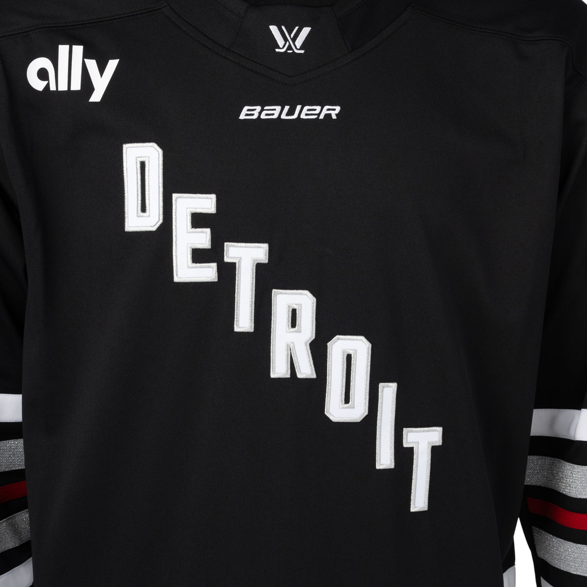 Front of a black jersey with red, white, and shimmering silver stripes on the sleeves. 'DETROIT' is in white with silver outlining diagonally across the chest. A white PWHL logo is on the front of the collar, a white Bauer logo below it, and a white Ally logo is on the right shoulder.