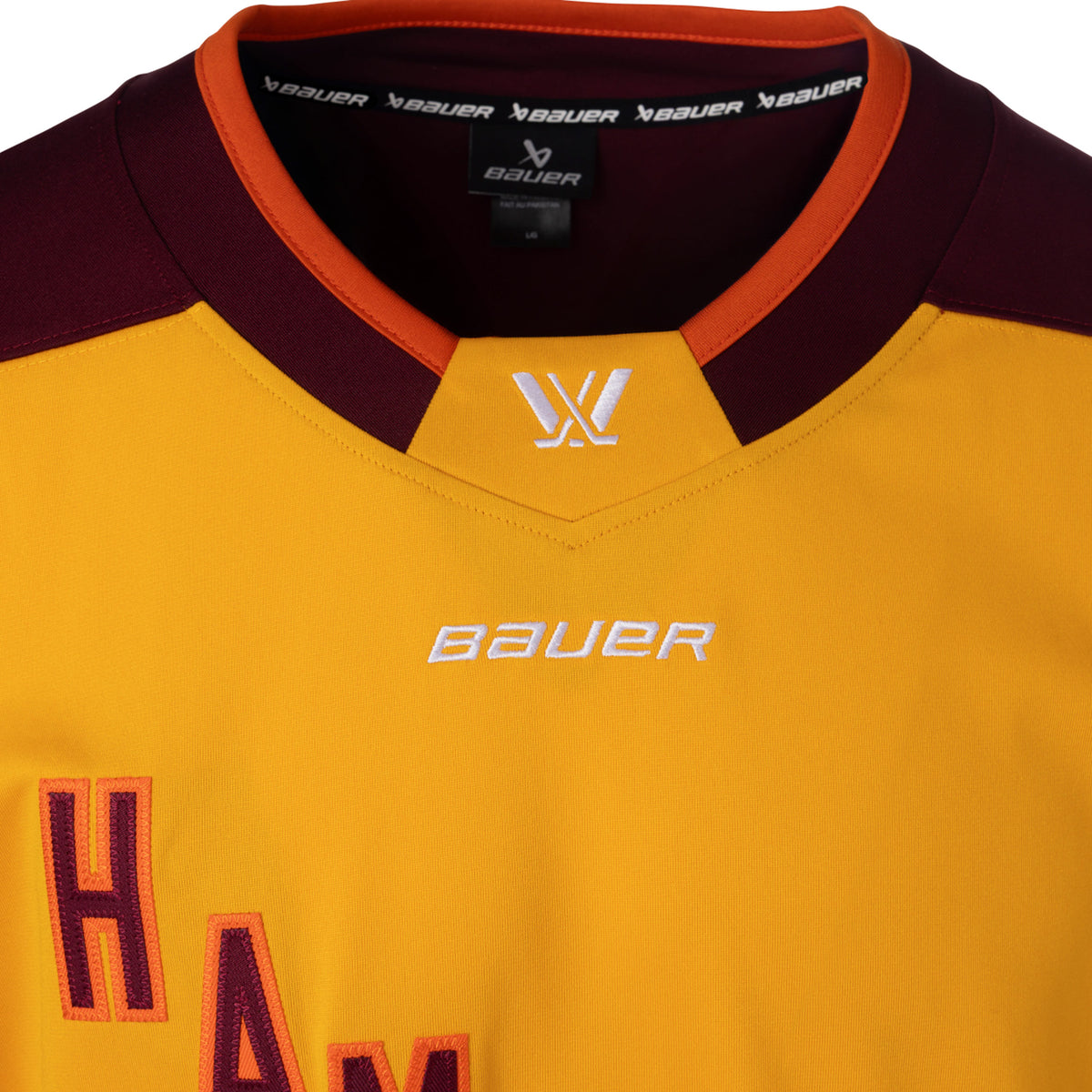 Front of a gold jersey with maroon shoulder caps. 'HAMILTON' is in maroon with orange outlining diagonally across the chest. The collar is maroon with orange trim and a gold color block in front showcasing a white PWHL logo with a Bauer logo below it. 