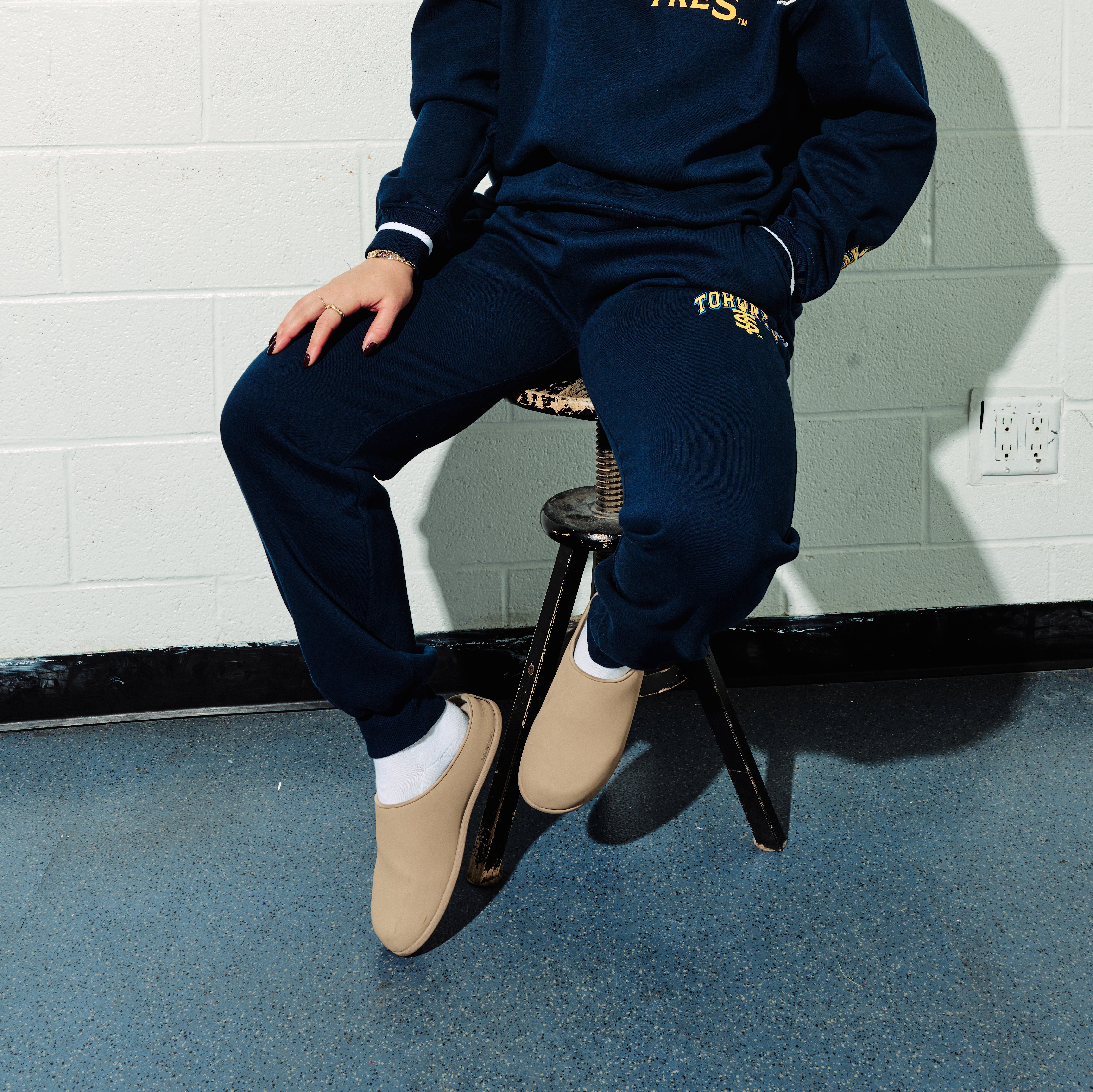 Person wearing navy tracksuit with yellow text, sitting on a stool against a tiled wall.