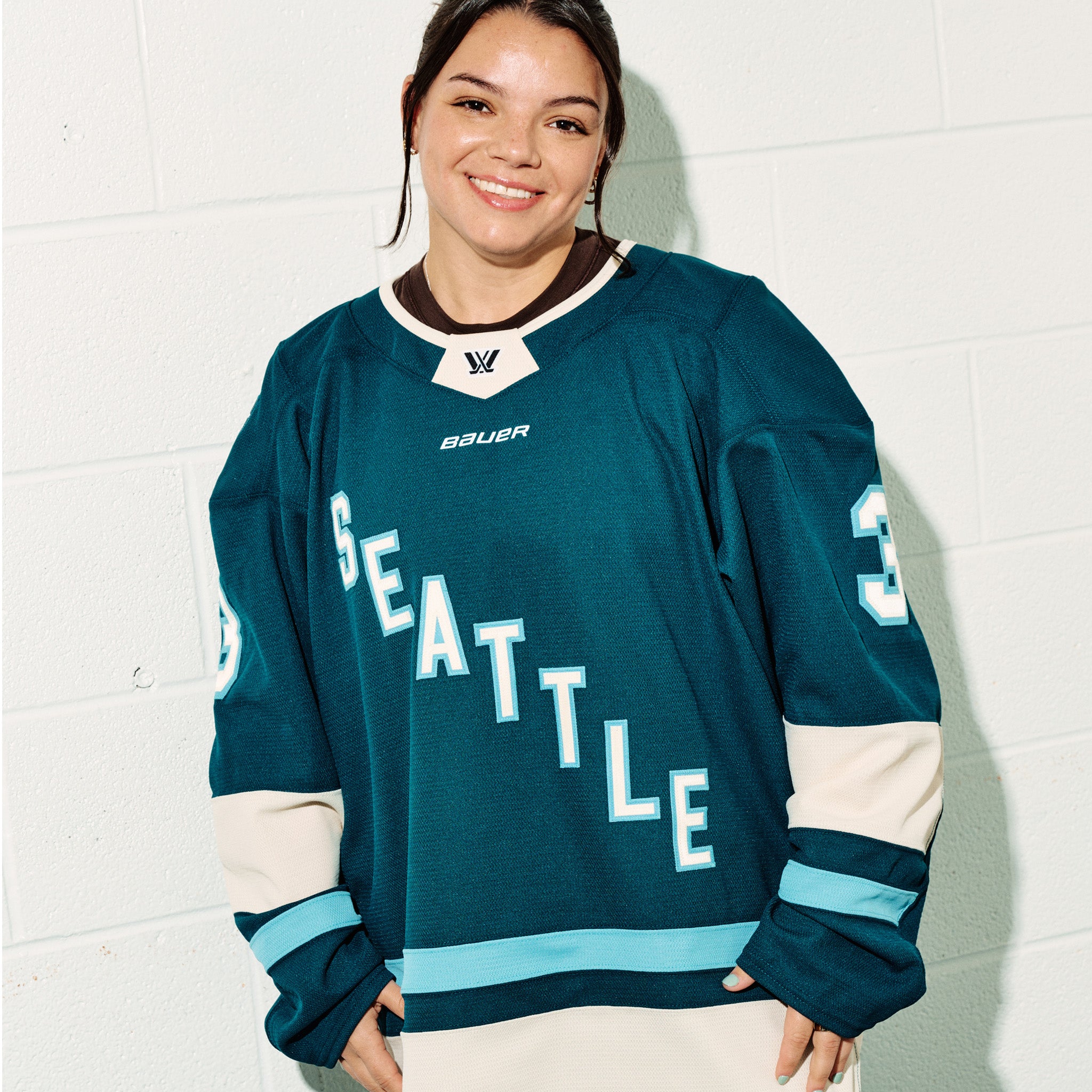 Cayla Barnes wearing her green PWHL Seattle jersey with, standing against a white tiled wall straight on angle.