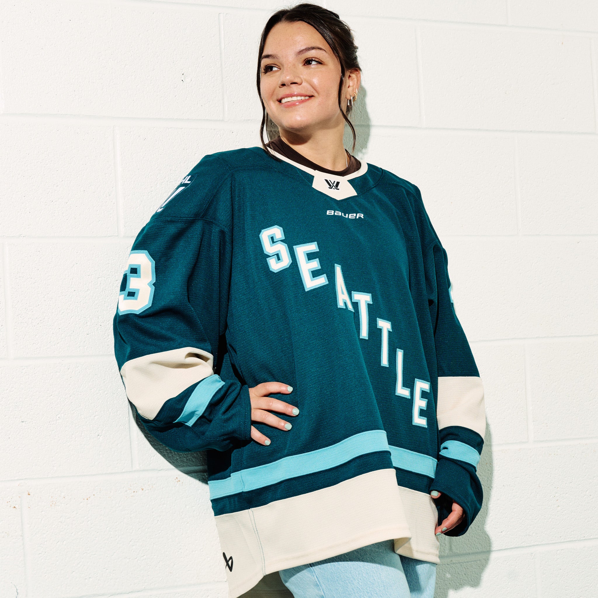 Cayla Barnes wearing her green PWHL Seattle jersey with, standing against a white tiled wall with her arm on her hip.