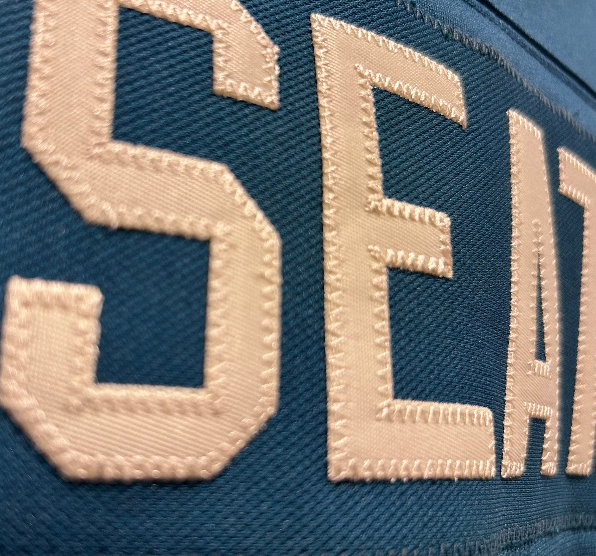 Close-up of a blue fabric with beige embroidered letters spelling 'SEATTLE'.