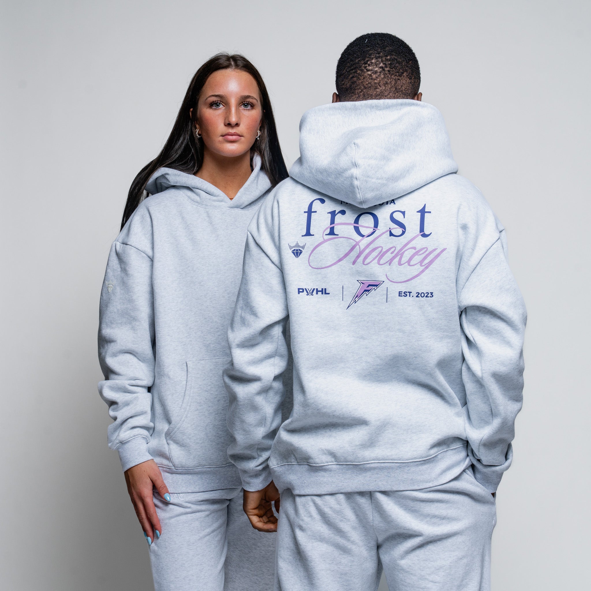 Two people wearing matching gray hoodies with branding on a plain background